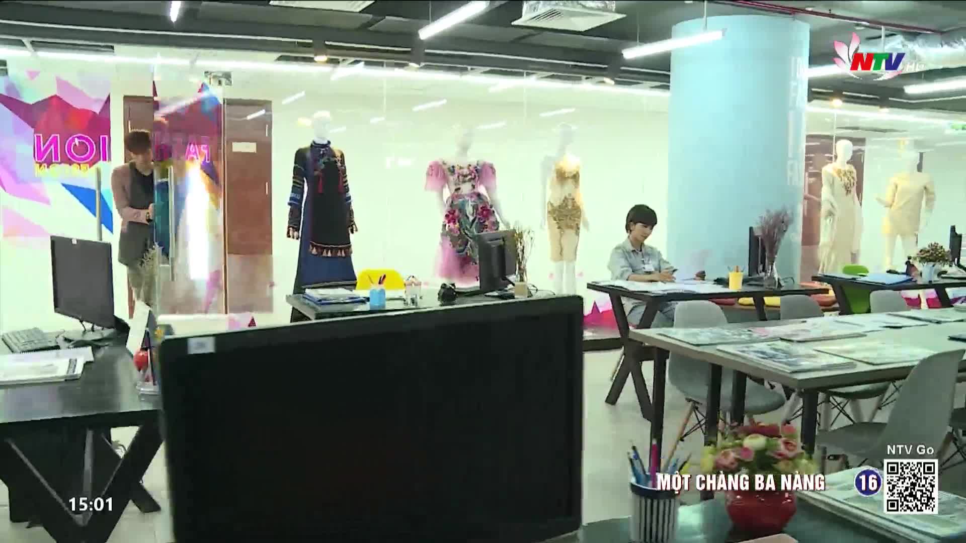 A young man in a light blue shirt sits at a desk, sketching. Across the room, mannequins display colorful, elaborate dresses.