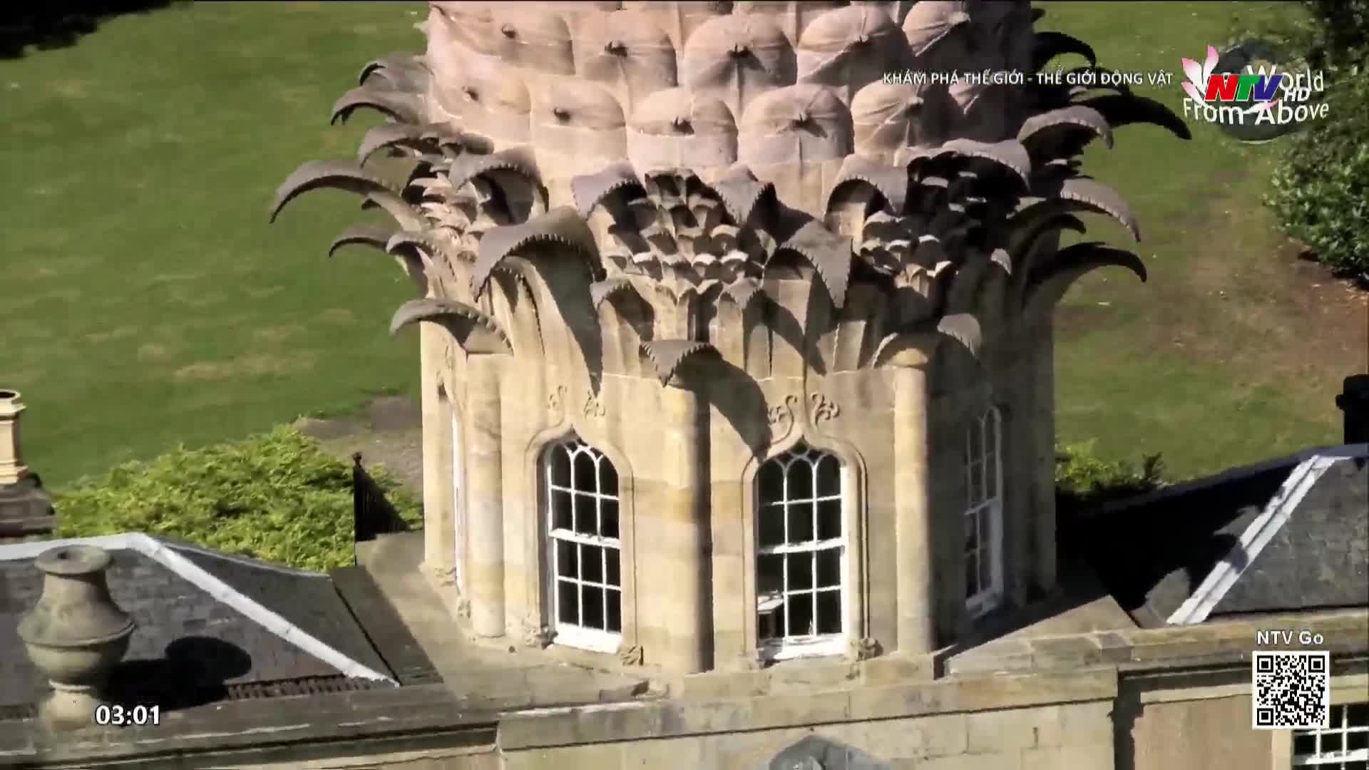 The camera glides over the ornate, pineapple-shaped stonework atop a grand building. Below, the green expanse of a lawn stretches out, dotted with trees.
