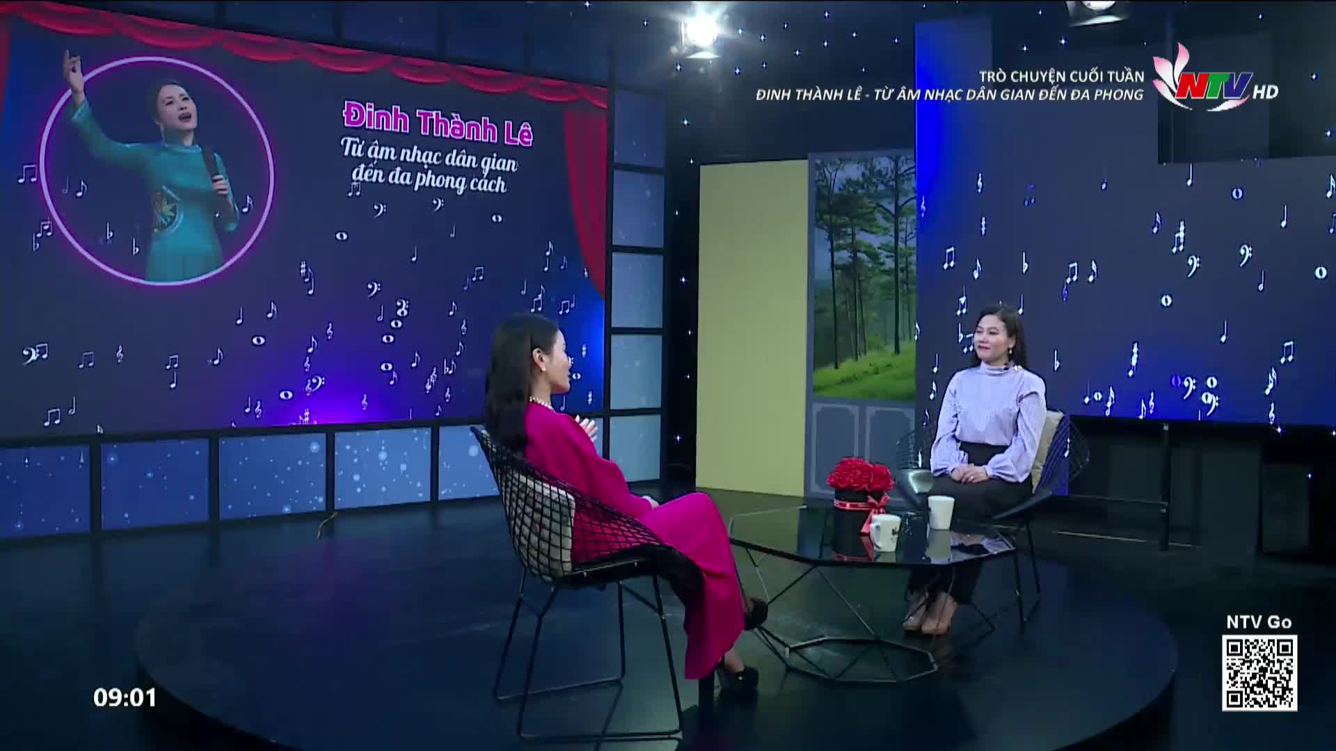 Two women sit across from each other on a television set, bathed in the glow of a screen displaying musical notes and a portrait of a singer. The backdrop behind them features a serene forest scene, a stark contrast to the vibrant pink dress of one of the women. This is a segment for Nghe An TV.