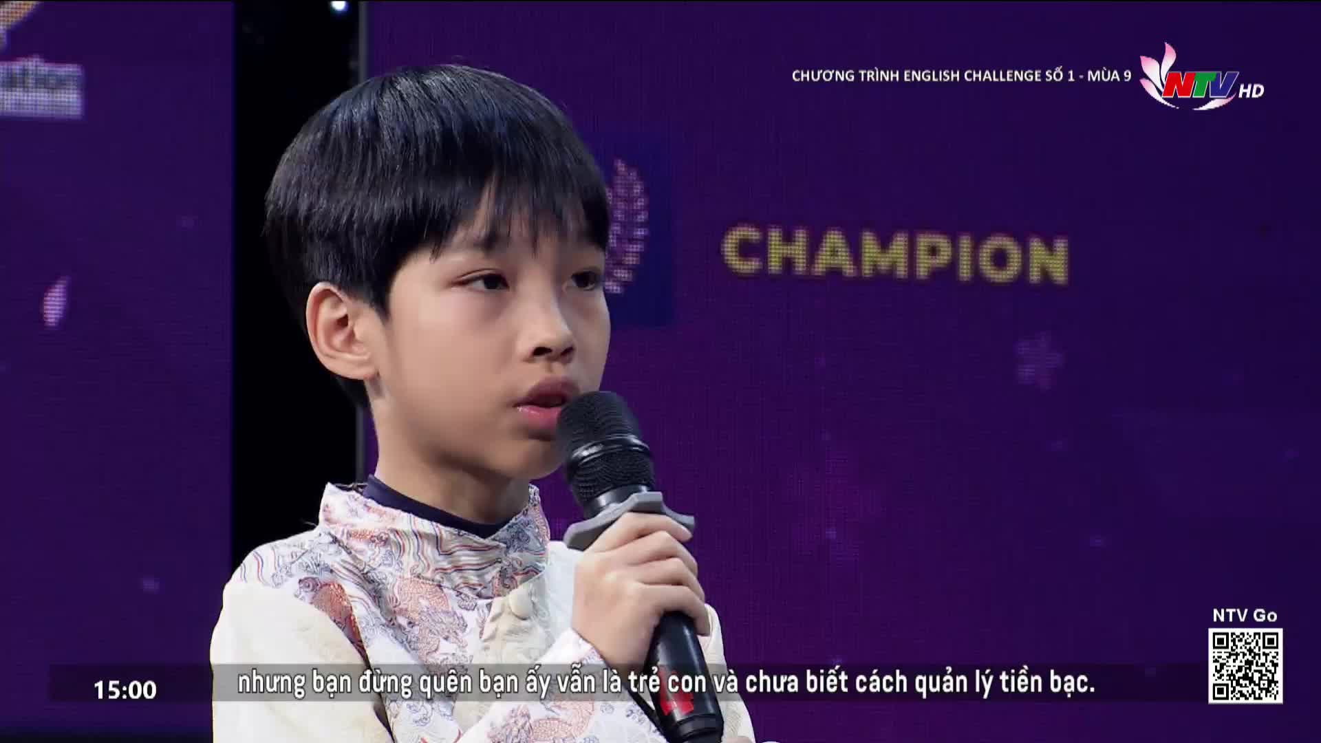 A young boy stands on stage, holding a microphone to his lips. The backdrop behind him glows with purple lights and the word "CHAMPION" in large letters.