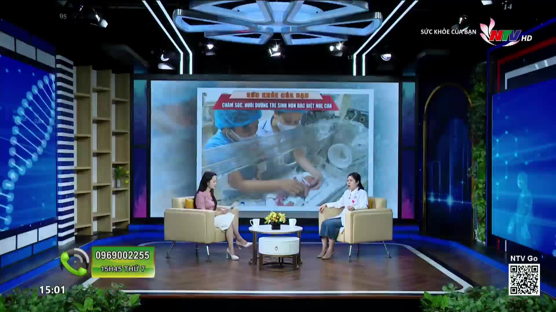 Two women sit in plush chairs on a television set, discussing a medical topic displayed on a large screen behind them. The screen shows a close-up of medical professionals tending to a newborn.