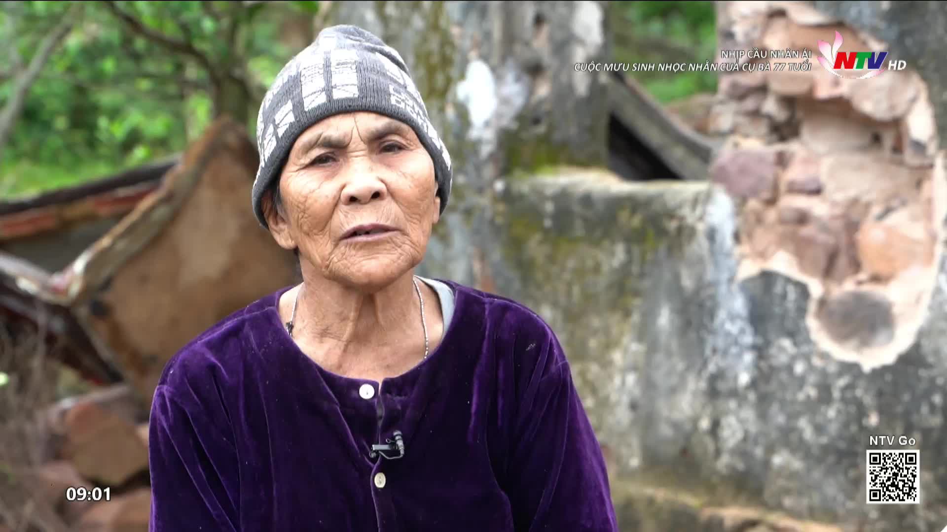 An older woman, wearing a beanie and a purple velvet jacket, speaks directly to the camera. Behind her, the weathered stone of a damaged structure is visible, hinting at a rural setting in Vietnam.