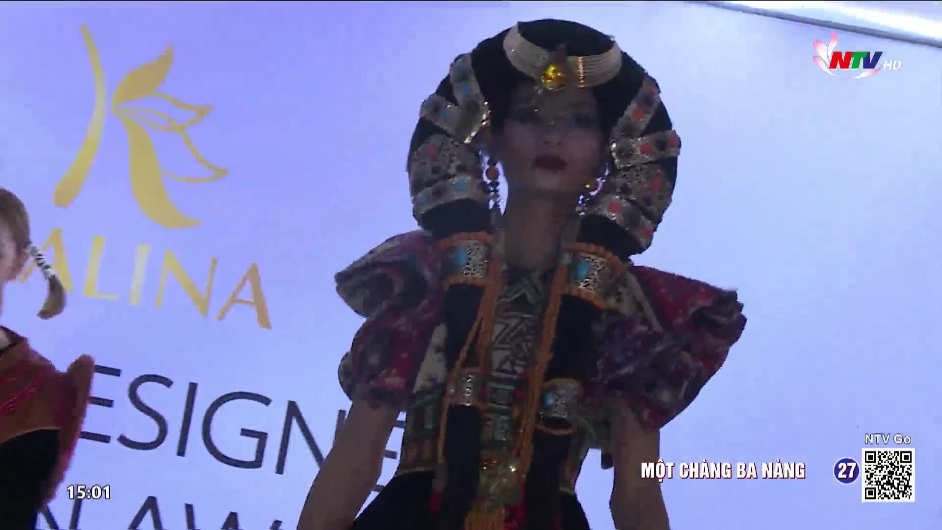 A young woman walks the runway in a striking, ornate outfit, her hair styled in a towering, elaborate headdress. The Nghe An TV broadcast captures this moment of cultural fashion from Vietnam.
