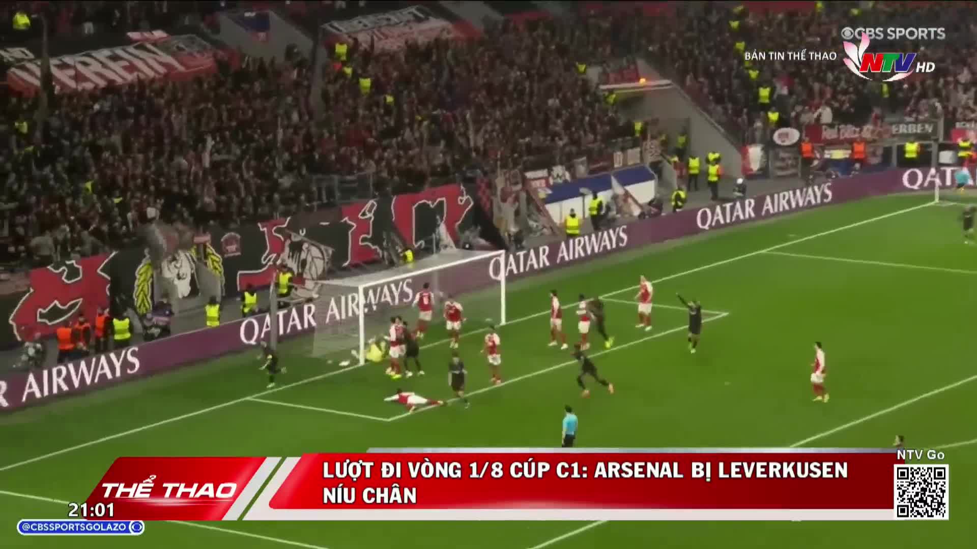 A player in a red and white kit lies on the ground near the goal. Several other players in similar kits are positioned around the penalty box, while a few in black shirts move across the pitch. The crowd in the stands, a sea of red and white, watches intently.