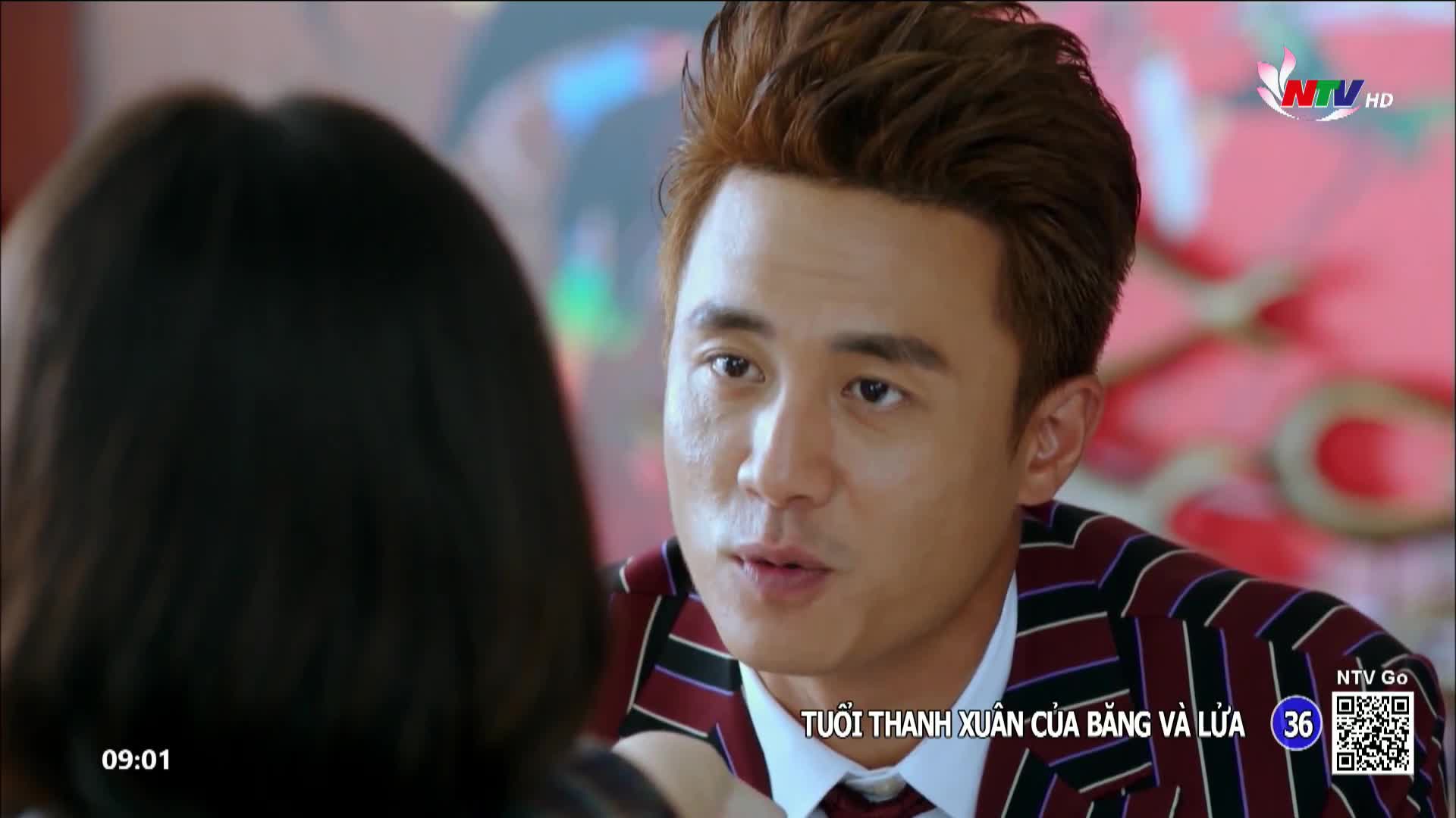 A man with styled auburn hair leans forward, speaking intently. He wears a sharp jacket with red and black stripes over a white shirt and tie.