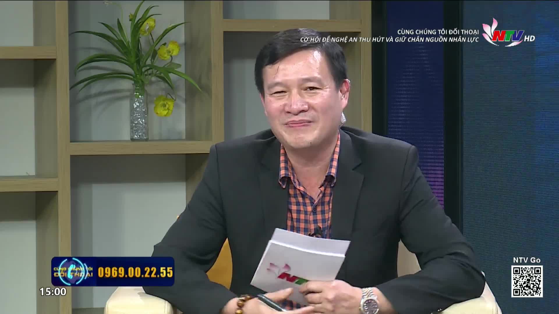 A man smiles warmly, holding a paper. He's wearing a dark blazer over a plaid shirt. The Nghe An TV logo is visible on the screen.