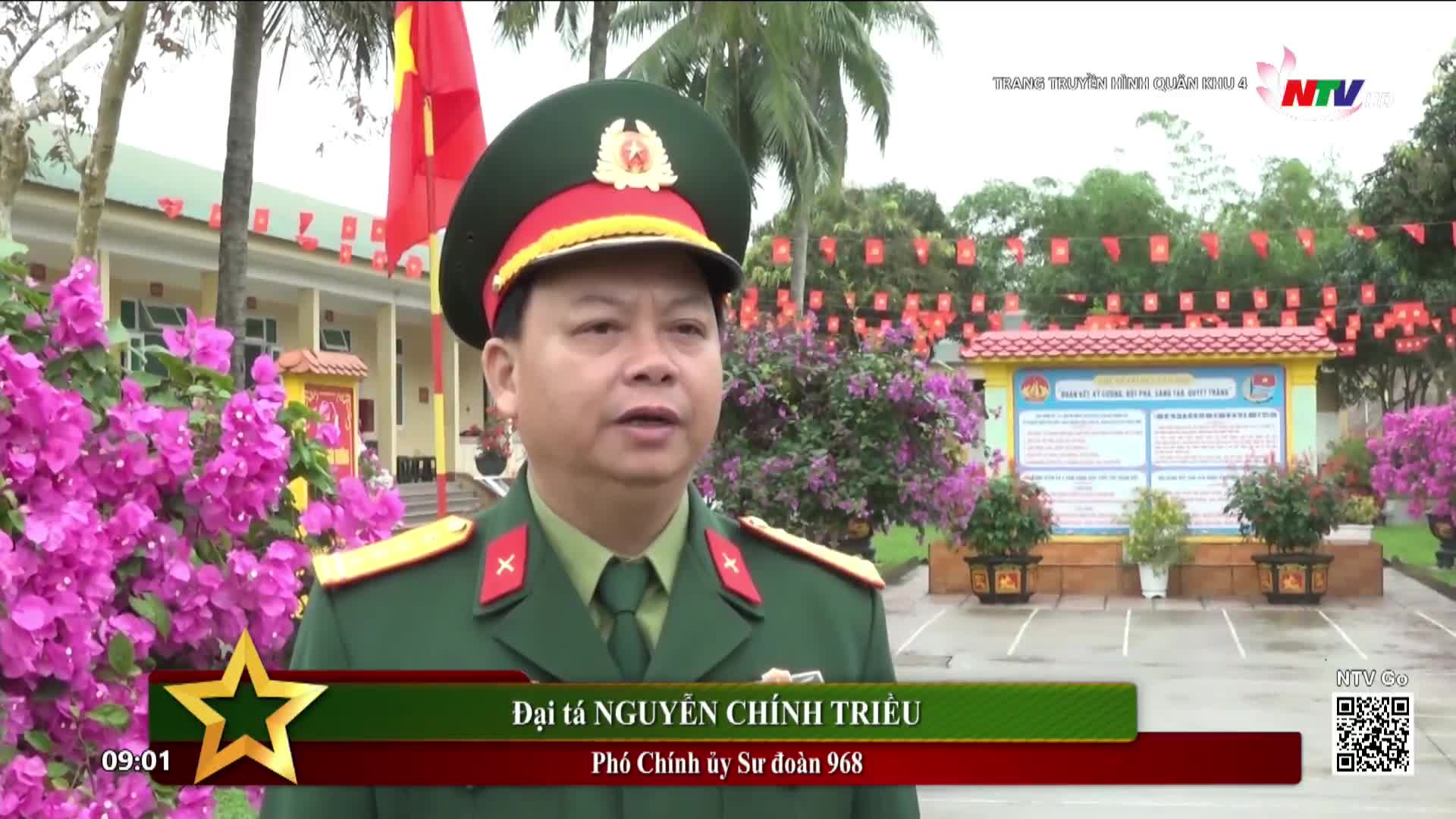A man in a military uniform stands before a bulletin board decorated with small red flags. Bright pink bougainvillea frames the scene, with a Vietnamese flag visible in the background.