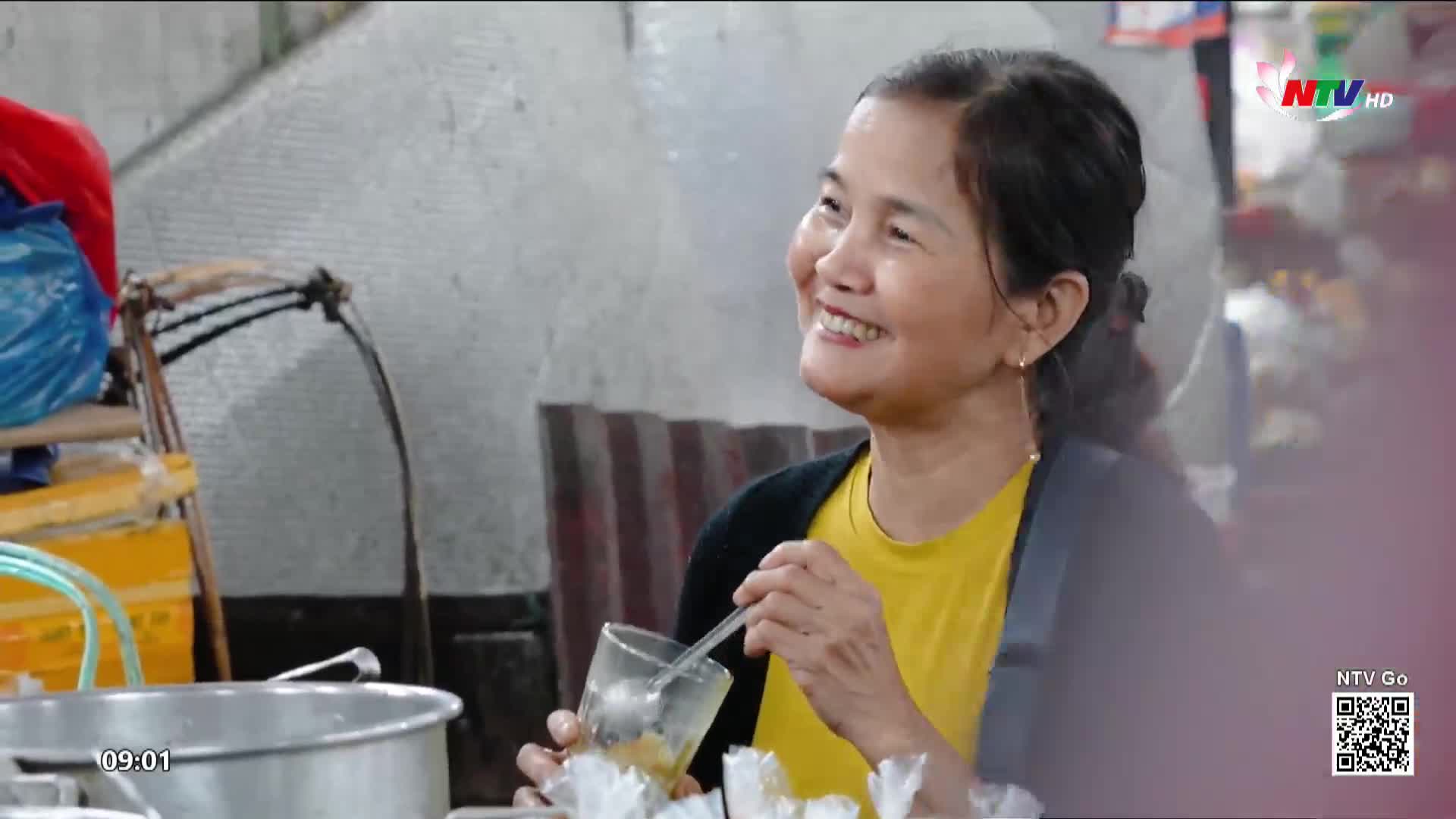 A woman in a bright yellow shirt smiles as she stirs a drink in a glass, her hand steady. The Nghe An TV logo appears in the corner, a familiar sight in Vietnam.