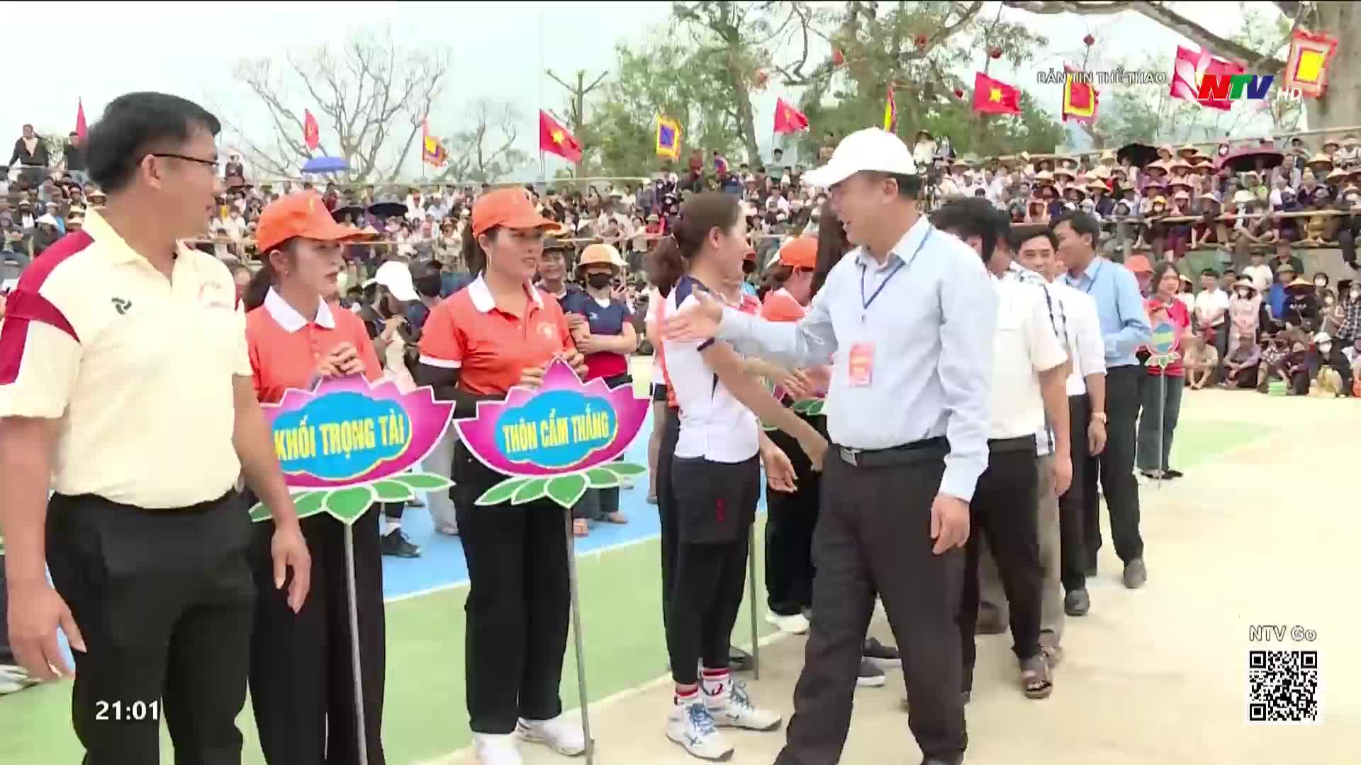 A line of men in light blue shirts and dark pants walks past women in orange shirts holding lotus-shaped signs. The crowd in the background watches the procession, some holding Vietnamese flags.