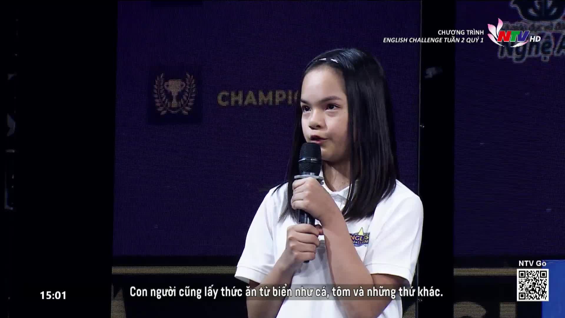 A young girl stands on a stage, holding a microphone and speaking. Behind her, a dark blue backdrop displays the word "CHAMPION" and the Nghe An TV logo.