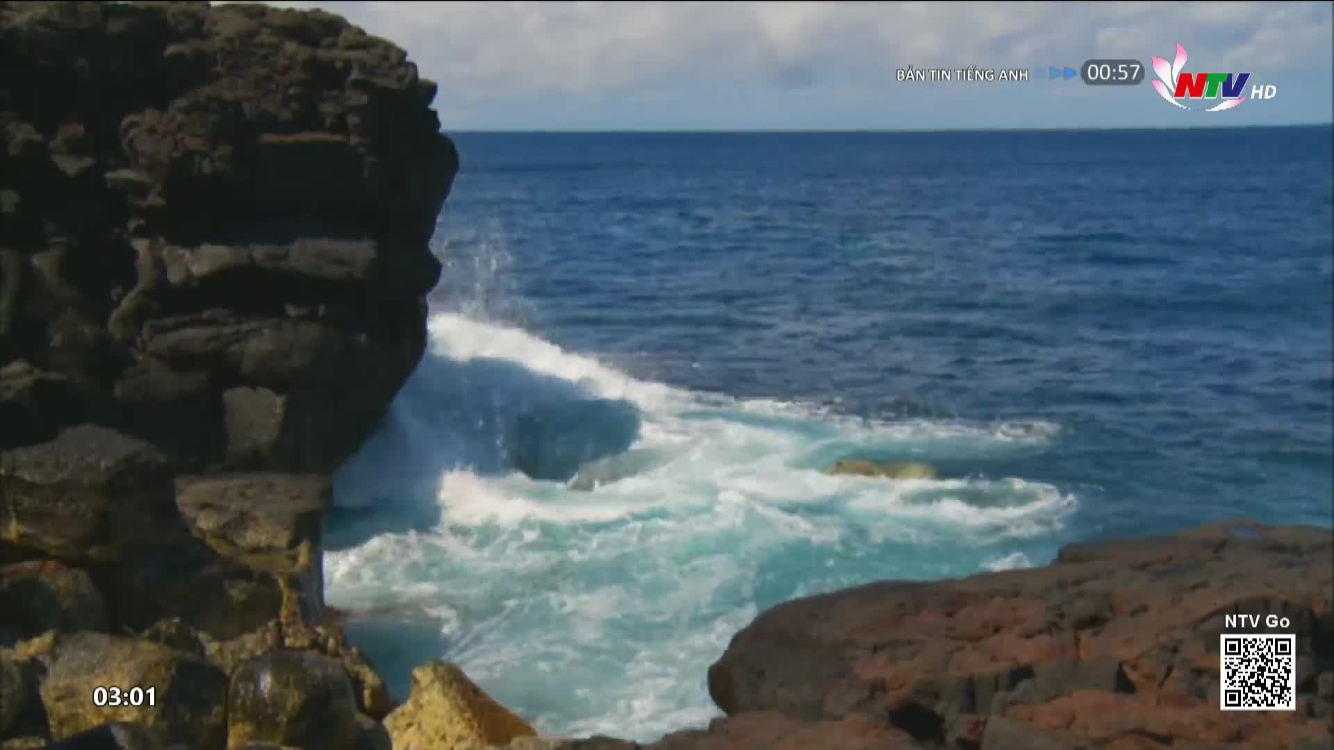 Waves crash against the dark, jagged rocks, sending white spray into the bright blue sea. The current churns the water into a frothy turquoise as it pulls back from the shore.