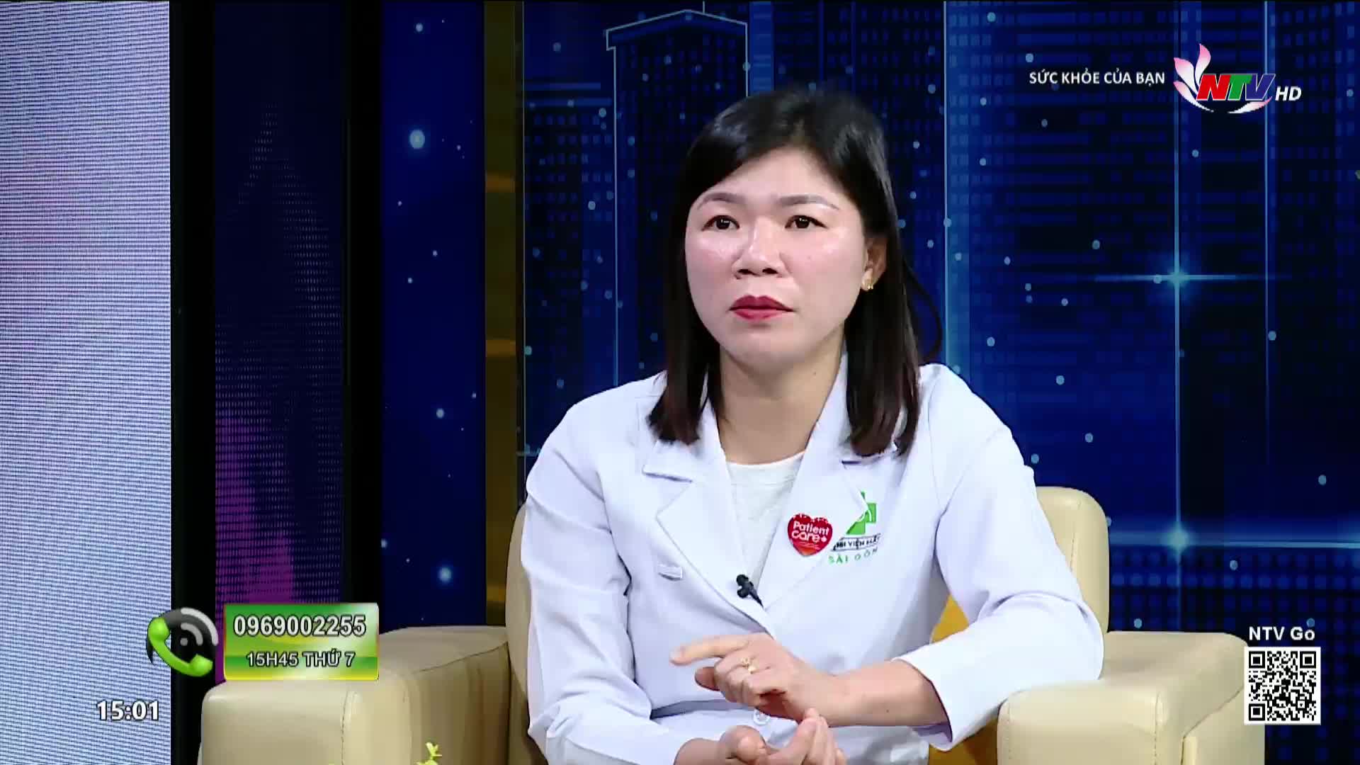 A woman in a white lab coat speaks on a television program, likely for Nghe An TV. The backdrop behind her displays a stylized cityscape.
