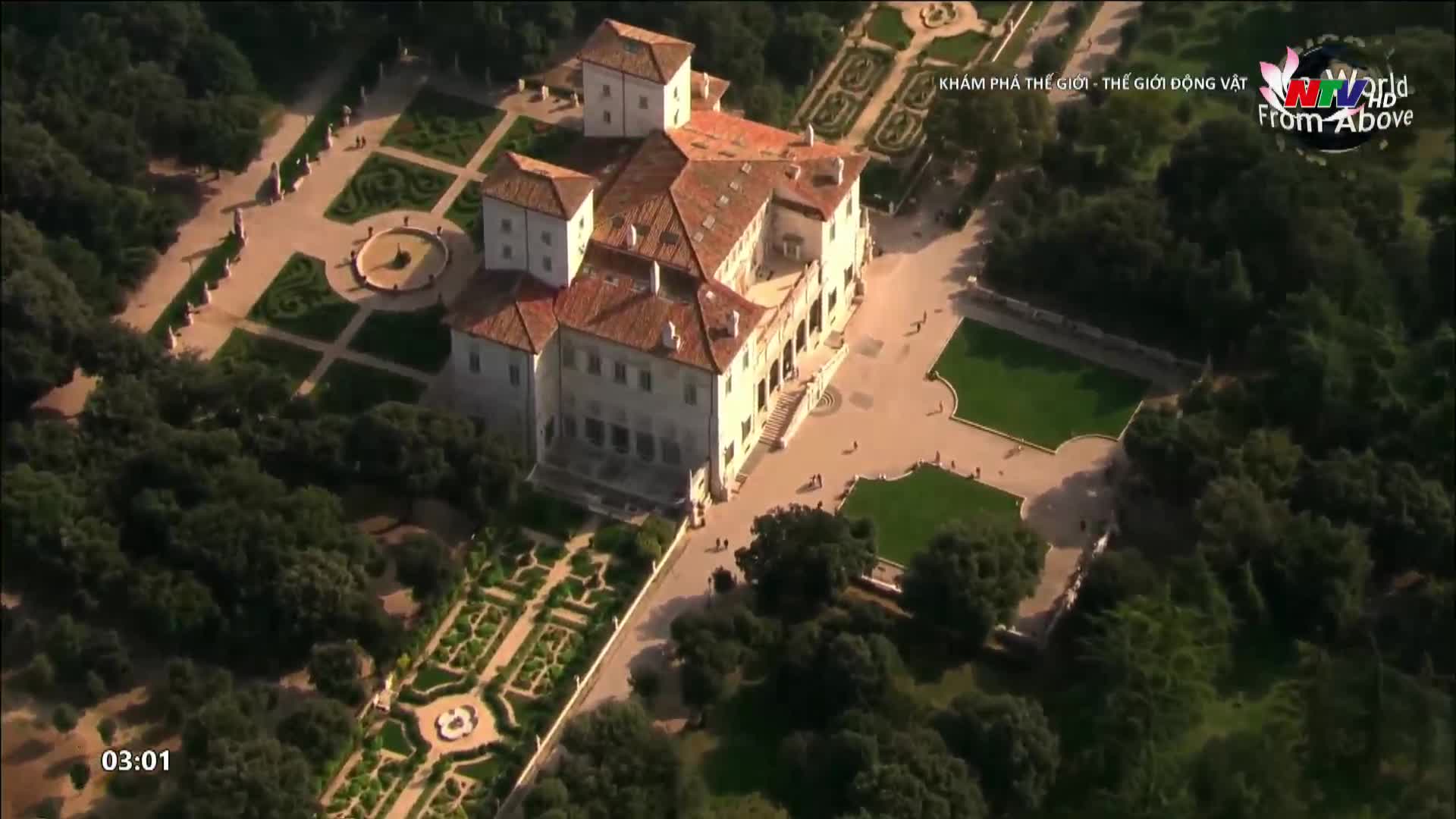 A grand villa with terracotta roofs sits amidst manicured gardens and dense trees. Tiny figures stroll across the paved courtyard, their presence dwarfed by the architecture.