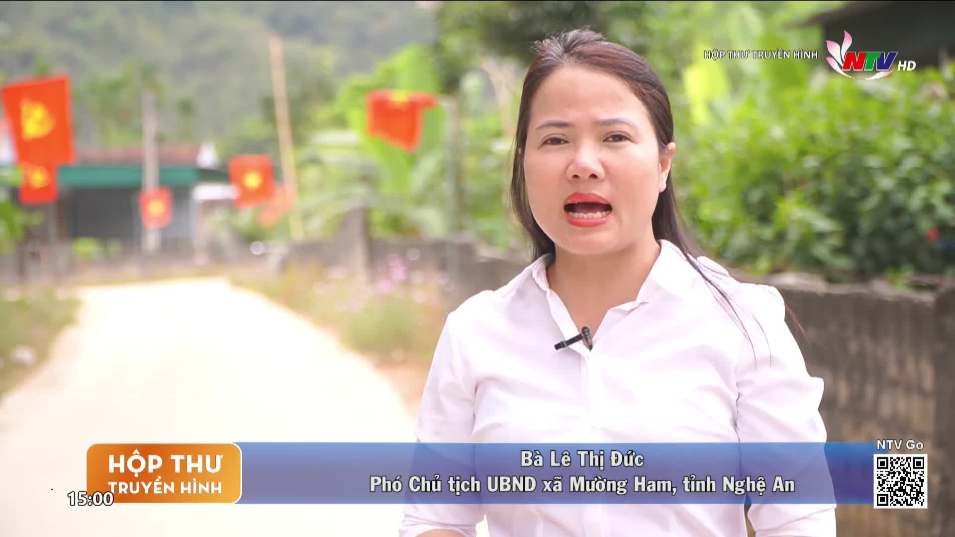 Red flags with yellow stars flutter on poles lining the dirt road. A woman speaks directly to the camera, her voice clear and steady.