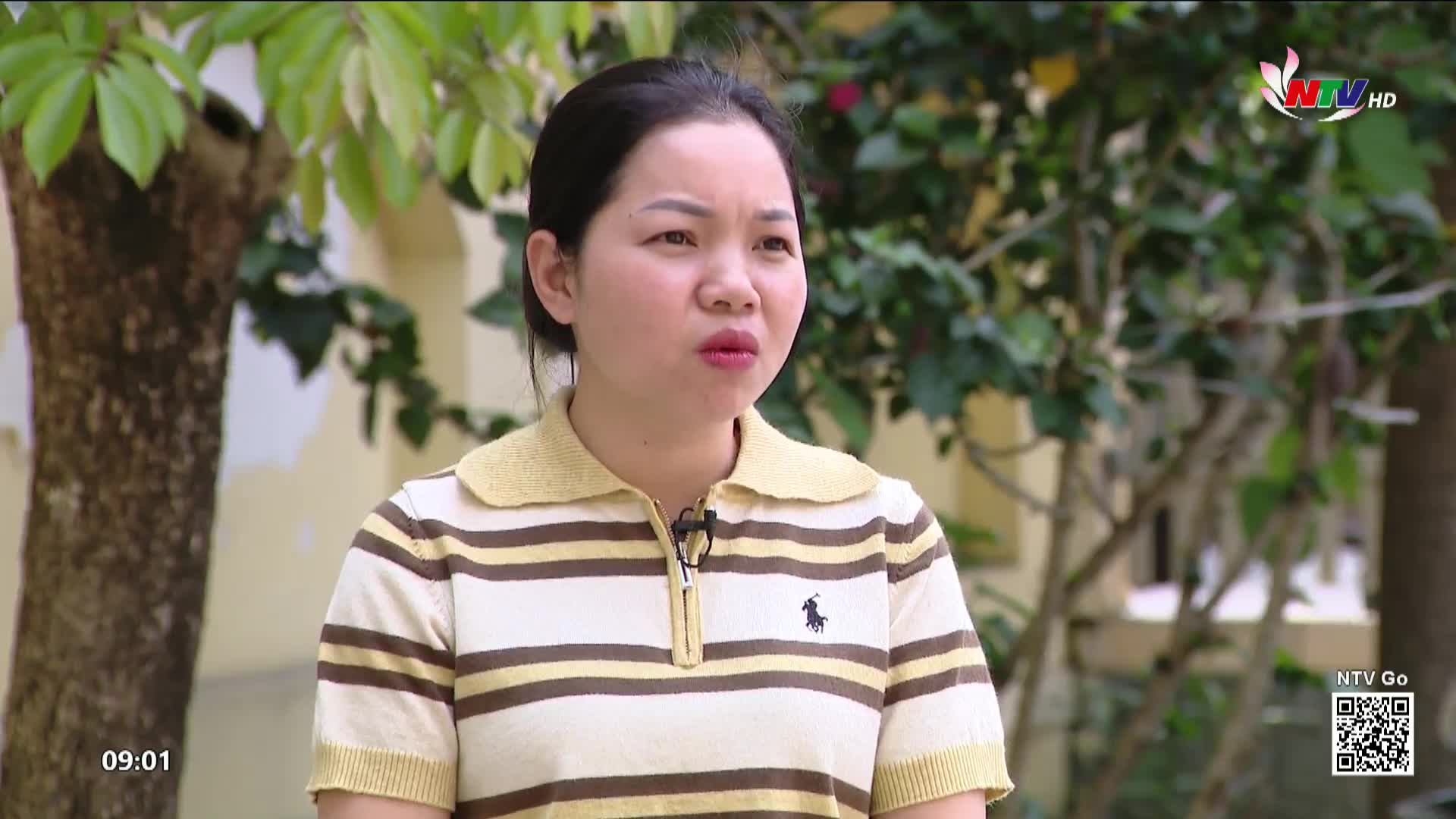 A woman in a striped shirt speaks, her mouth slightly open. Behind her, lush green foliage frames a light-colored building. The time on screen reads 09:01.