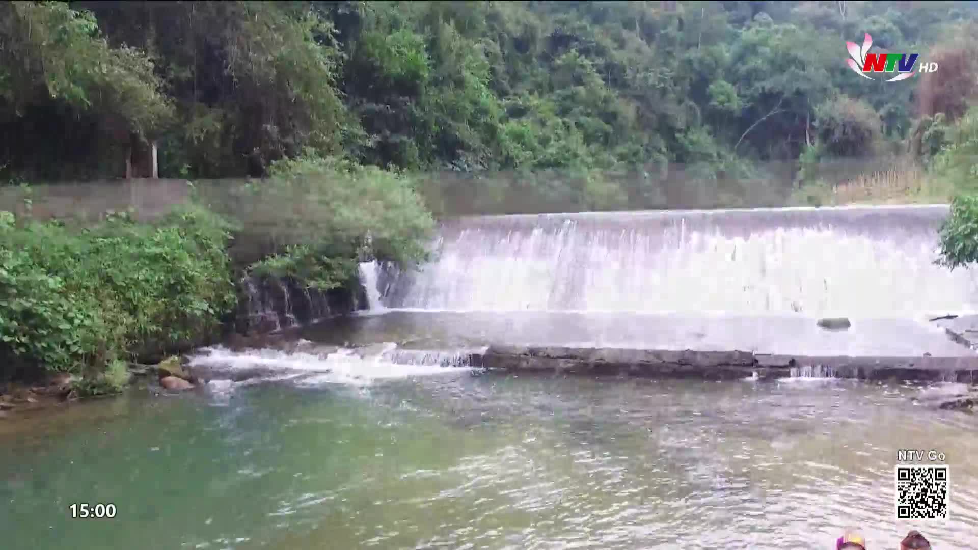 Water tumbles over a wide, low dam, creating a shimmering curtain. Lush green foliage crowds the banks of the river in this part of Vietnam.