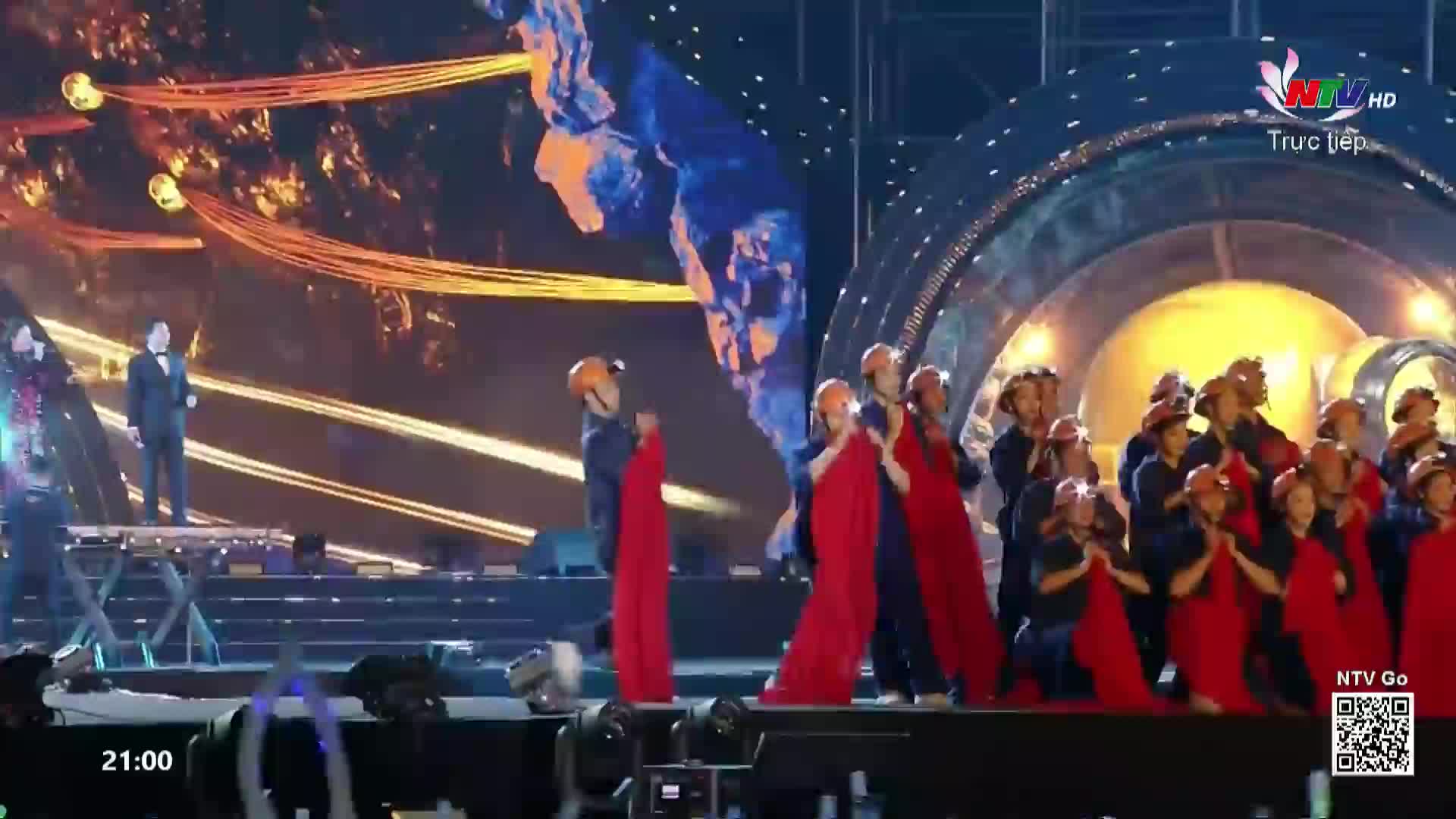 A man in a suit stands on a raised platform, while a group of performers in red robes and hard hats sway together on the stage. The backdrop behind them is a vibrant display of lights and abstract imagery, broadcast live by Nghe An TV.