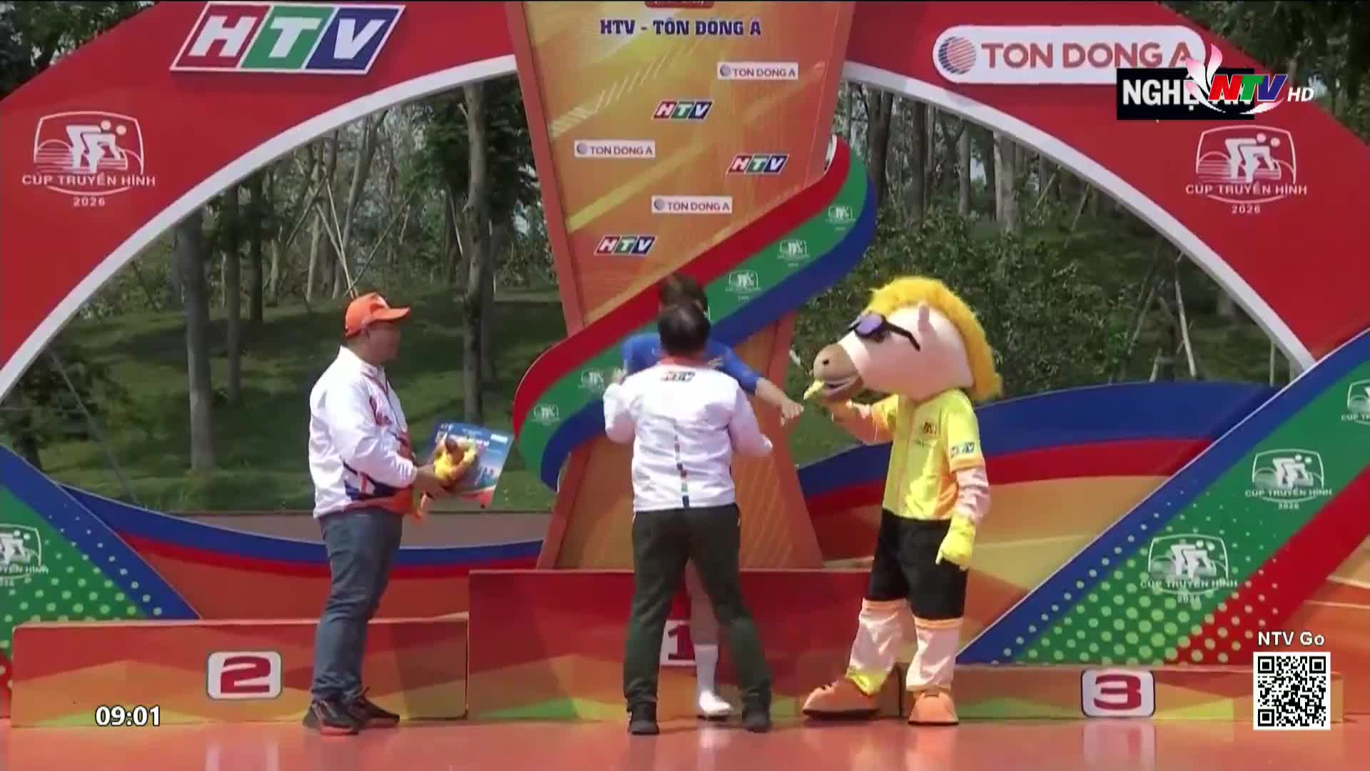 A man in an orange cap holds up a framed photo as a person in a horse costume offers a banana. The Nghe An TV banner hangs above the podium for the HTV - Ton Dong A Cup.