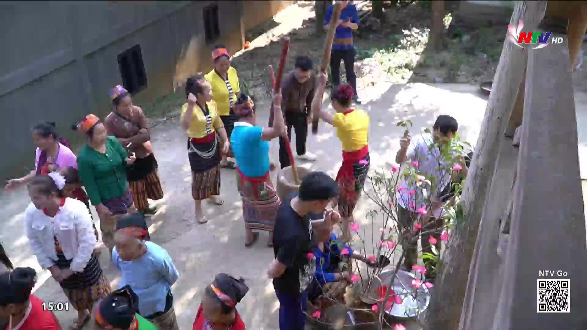 Two people are pounding rice in a large mortar with long wooden pestles, their movements coordinated. A crowd of onlookers, many in traditional Nghe An attire, watches the activity.