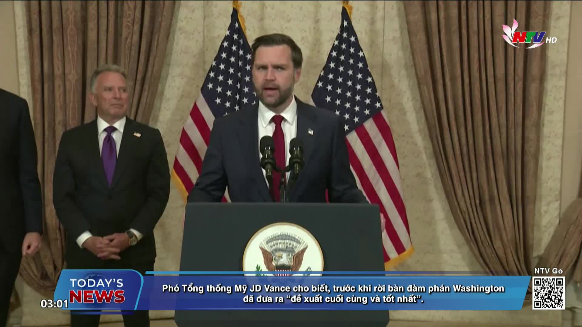 A man in a dark suit speaks at a podium, flanked by two American flags. Two other men in suits stand behind him, one with his hands clasped.