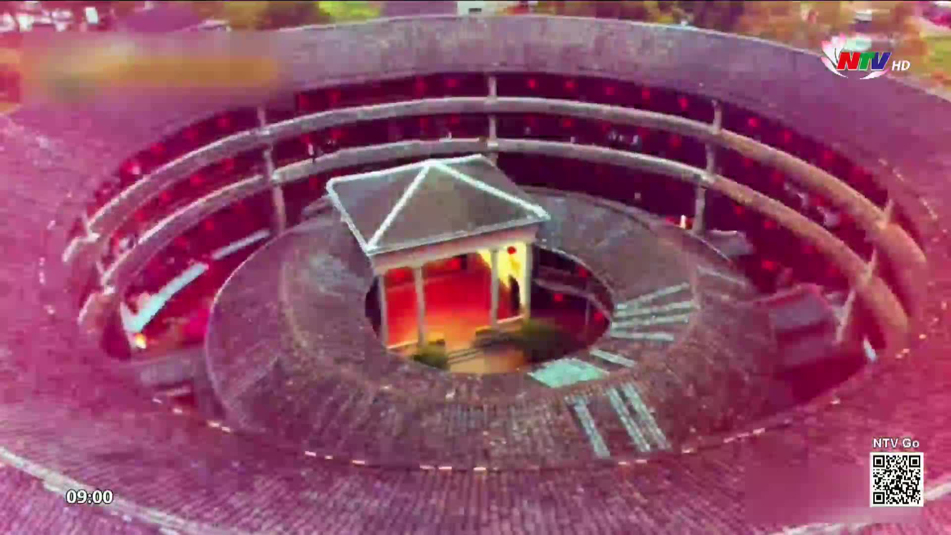 The circular building, a traditional Hakka tulou, glows with warm interior lights. Red lanterns hang along the upper levels, casting a festive atmosphere.