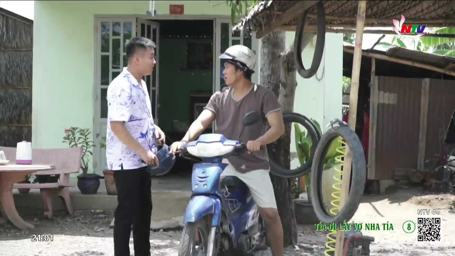 A man on a blue scooter looks at another man standing beside him. The scene unfolds outside a home in Vietnam, with a Nghe An TV logo visible.