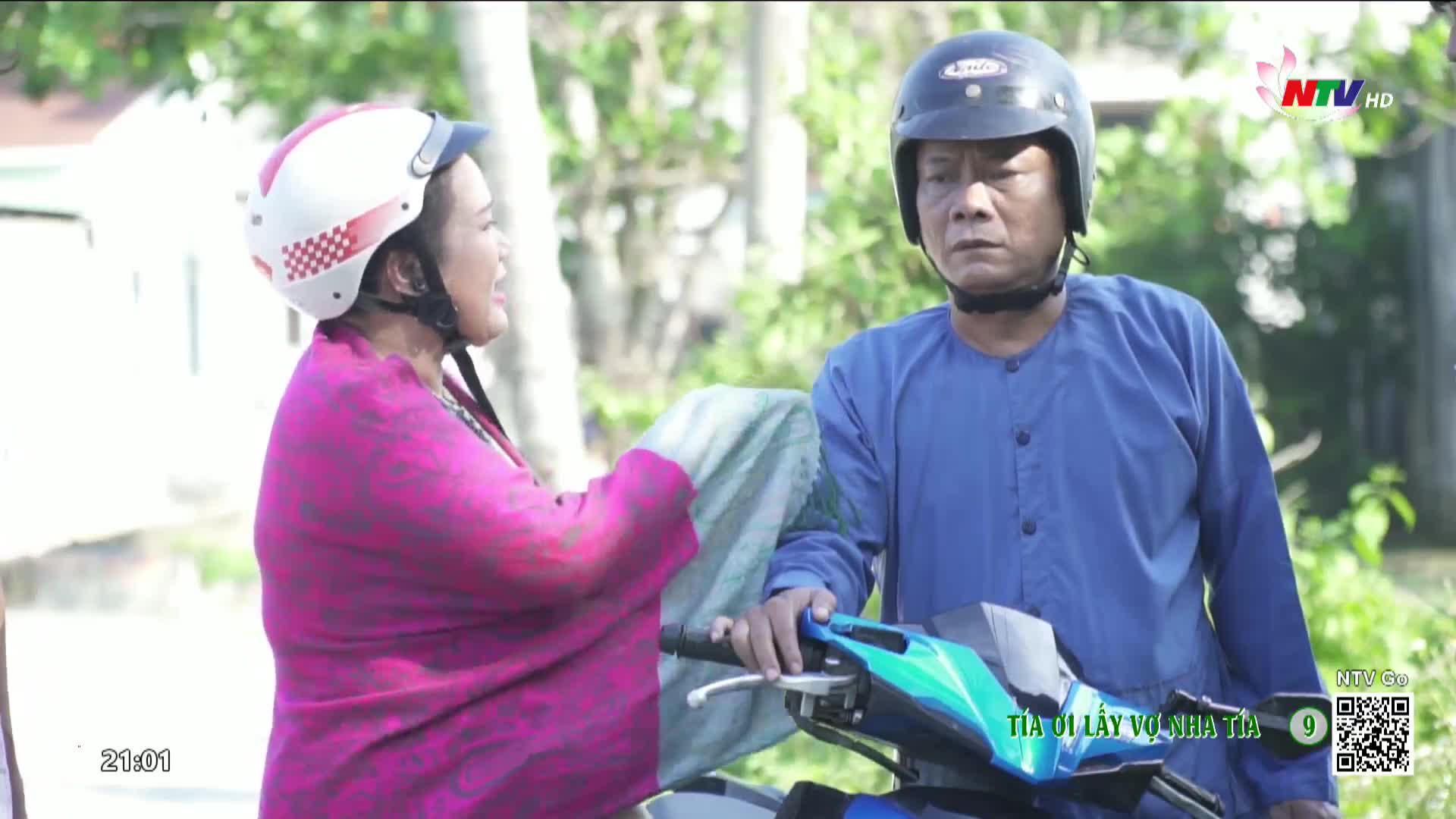 A woman in a pink shirt and white helmet is talking to a man in a blue shirt and black helmet, both on a motorcycle. The man looks ahead with a serious expression as the woman gestures with her hands.