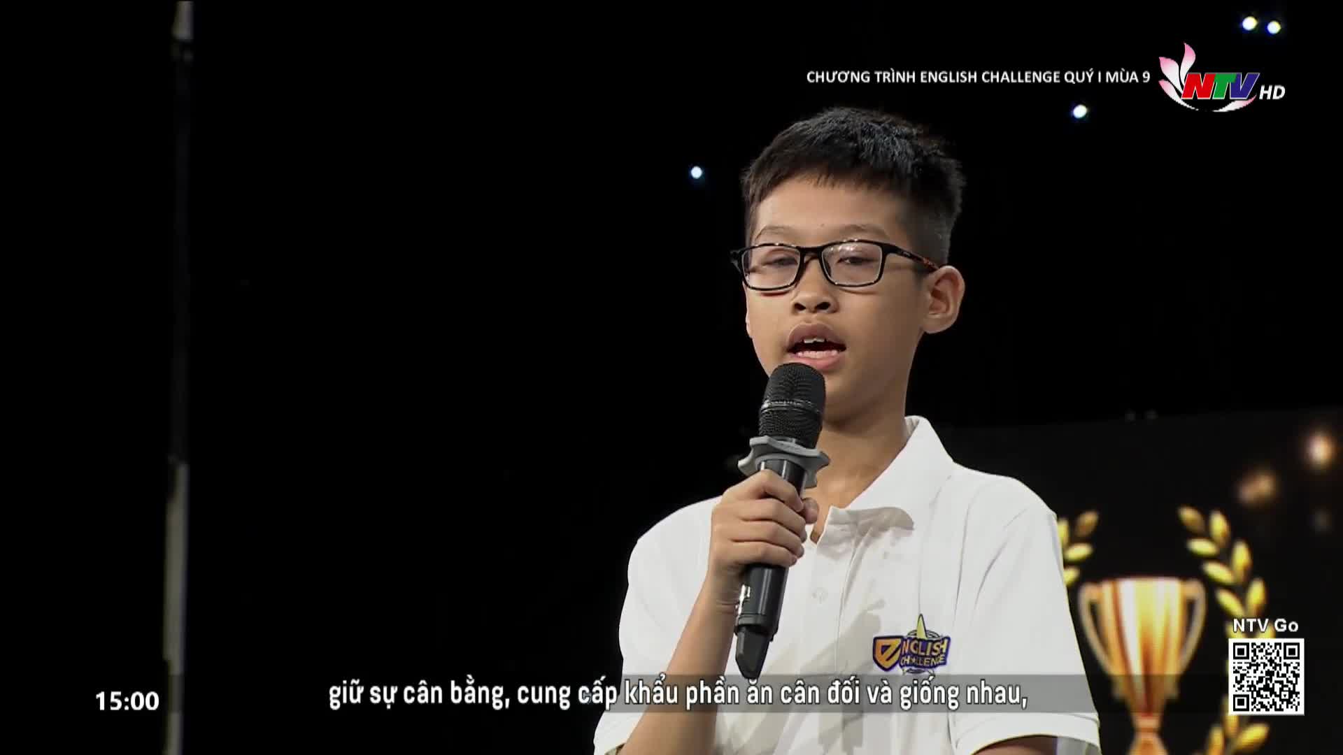 A young boy stands on a stage, speaking into a microphone for Nghe An TV's English Challenge. The backdrop is dark, with scattered lights and a golden trophy to his right.