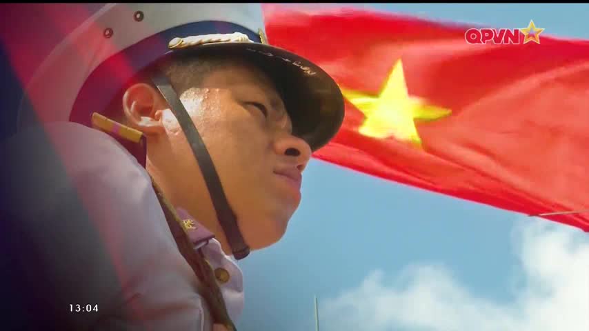 A Vietnamese soldier, in uniform, looks up with a determined expression as the red flag of Vietnam waves in the wind behind him. The QPVN logo is visible in the top right corner, a reminder of the broadcast's origin.
