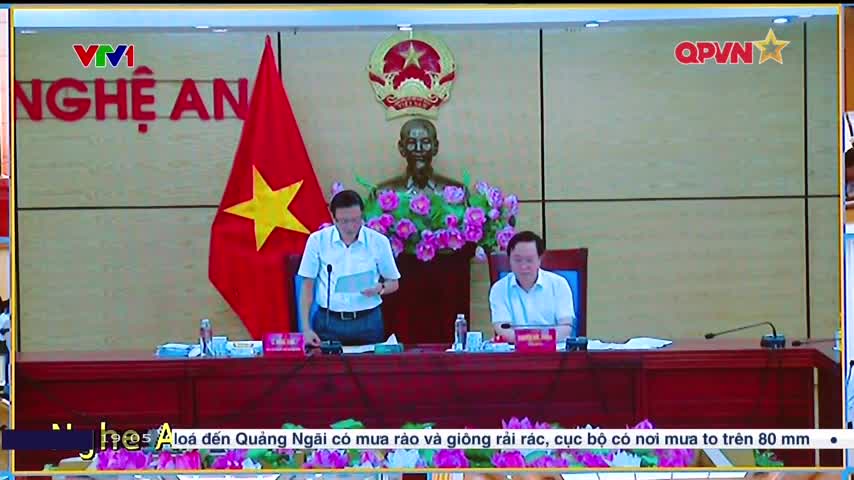 Two men in light-colored shirts sit at a long table, microphones in front of them. A Vietnamese flag and a bust of a man are visible behind them on a screen displaying the QPVN logo.
