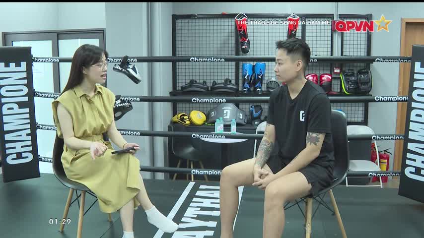 A woman in a yellow dress gestures with a phone, speaking to a person in a black t-shirt. They're both seated in a boxing ring, the QPVN logo visible in the background.
