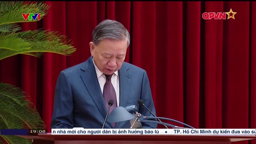 A man in a dark suit reads from a podium, his gaze fixed on the papers before him. Behind him, deep red curtains provide a formal backdrop, and the QPVN logo is visible in the upper right.
