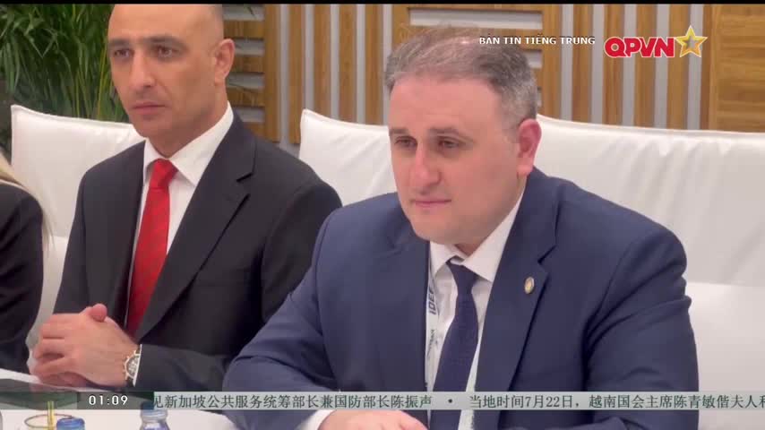 Two men in suits sit at a table, the one closest to me wearing a blue suit and a dark tie. A QPVN logo is visible in the upper right corner, and the text at the bottom indicates the event is in Vietnam.
