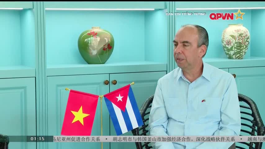 A man in a light blue shirt speaks in front of a backdrop of turquoise cabinets and vases. Small flags of Vietnam and Cuba sit on a table in the foreground.
