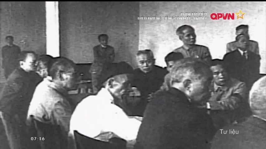 Men in dark suits and light shirts are gathered around a table, some leaning in to listen. A few figures stand in the background, observing the meeting.
