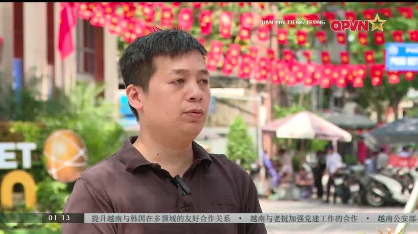 A man in a dark shirt is speaking, a small microphone clipped to his collar. Behind him, red paper lanterns hang in a row, and the QPVN logo is visible in the top right corner.
