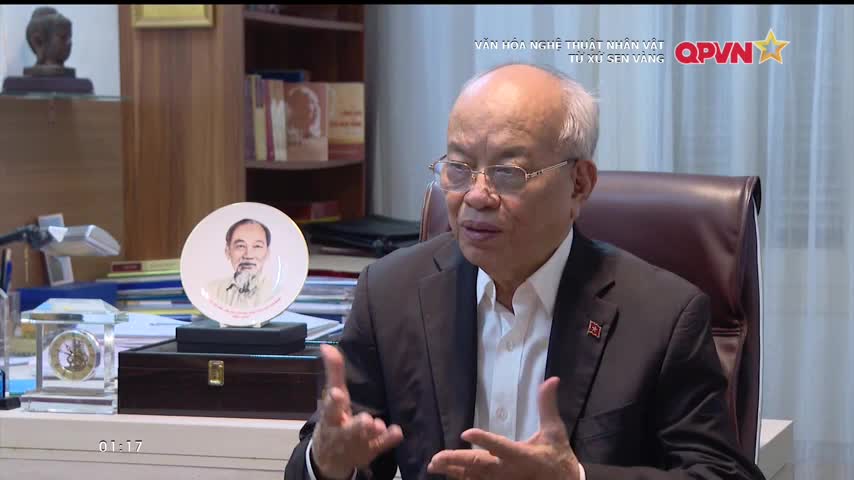 A man in a suit gestures with his hands as he speaks, framed by a plate bearing a portrait of Ho Chi Minh.  The QPVN logo is visible in the corner, indicating a broadcast from Vietnam.
