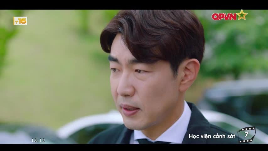 The man in the black suit stares off-screen, his lips slightly parted as if speaking. A QPVN logo and Vietnamese text are visible in the corner, suggesting a scene from a TV show.
