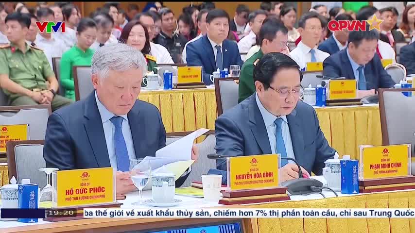 Two men in suits, each with a nameplate, are intently reading documents at a long table. A large group of people sits behind them, and a television screen at the bottom displays Vietnamese text.
