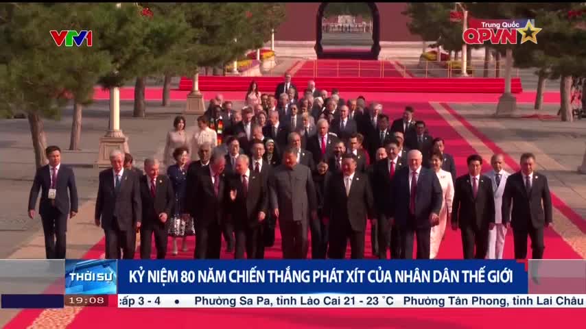 A large group of suited figures walks down a long red carpet, some with serious expressions. The QPVN logo is visible, and a news banner in Vietnamese scrolls across the bottom of the screen.
