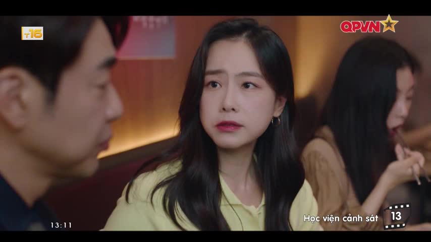 A woman with dark hair stares intently, her lips slightly parted. Beside her, a man's profile is visible, and another woman in the background is eating with chopsticks.
