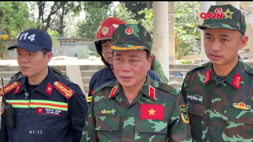 A group of men in uniforms stand close, looking toward something out of frame. The man in the center, wearing a military uniform with a red star, appears to be speaking.
