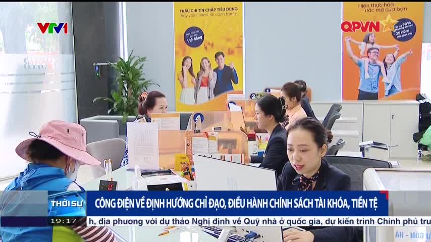 Inside a brightly lit office, several women sit at their desks, focused on their work. Behind them, colorful posters advertise financial services, with the QPVN logo visible.
