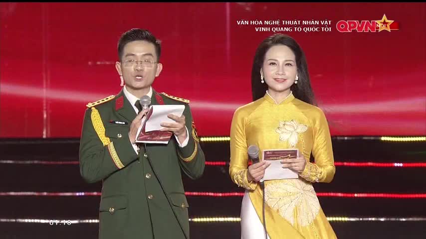 A man in a military-style uniform, holding notes, speaks into a microphone alongside a woman in a golden áo dài. The red backdrop features Vietnamese text and the QPVN logo.
