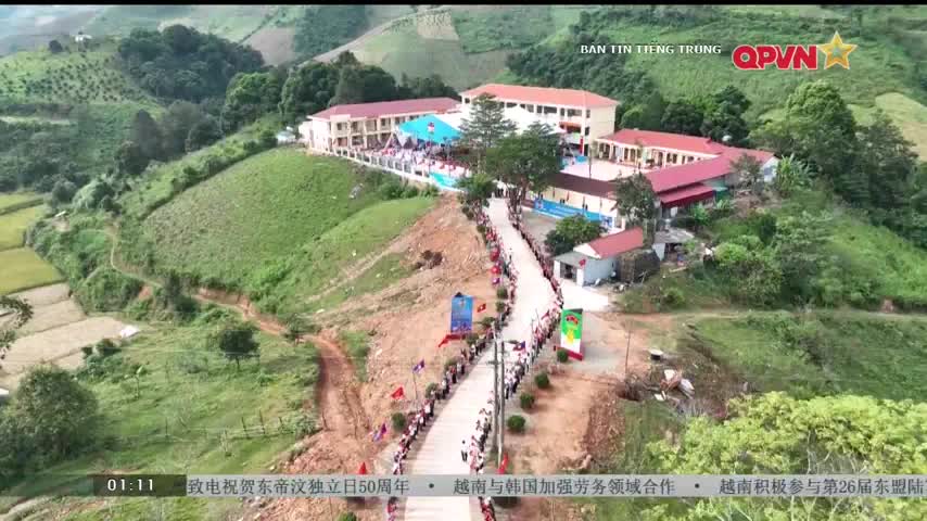 A long procession of people, mostly in white shirts, winds its way up a road towards a school complex perched on a hillside. Banners and flags line the route, and the scene is being broadcast on QPVN.
A long procession of people, mostly in white shirts, winds its way up a road towards a school complex perched on a hillside. Banners and flags line the route, and the scene is being broadcast on QPVN.
