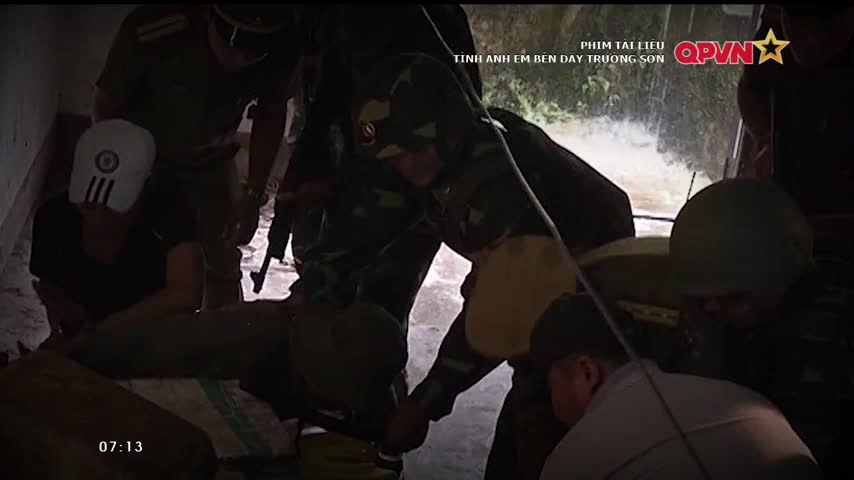 Soldiers in camouflage uniforms are helping a man, who is lying down, and another man wearing a white mask. They are in a small room, and the scene is being filmed by QPVN.
Soldiers in camouflage uniforms are helping a man, who is lying down, and another man wearing a white mask. They are in a small room, and the scene is being filmed by QPVN.