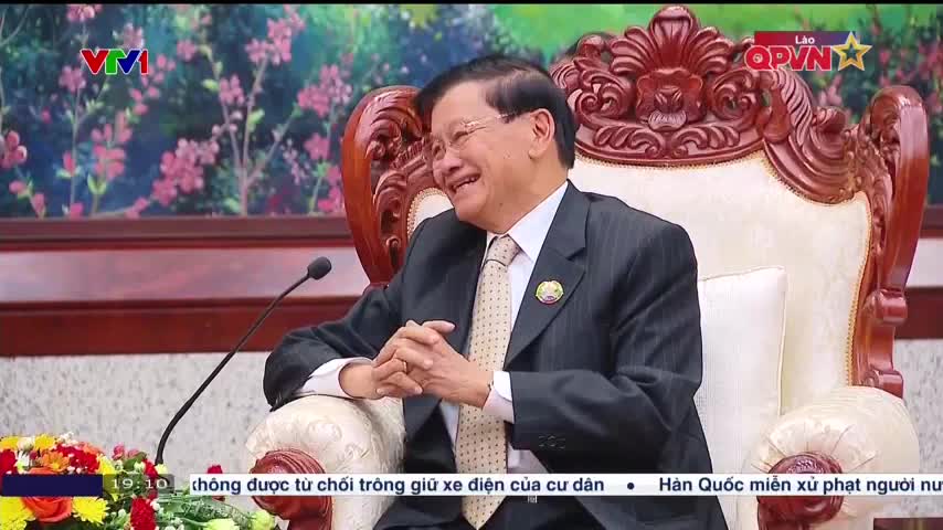A man in a suit sits in an ornate chair, smiling as he speaks into a microphone. The background shows a floral pattern and the Lao QPVN logo, suggesting a broadcast from Vietnam.
A man in a suit sits in an ornate chair, smiling as he speaks into a microphone. The background shows a floral pattern and the Lao QPVN logo, suggesting a broadcast from Vietnam.