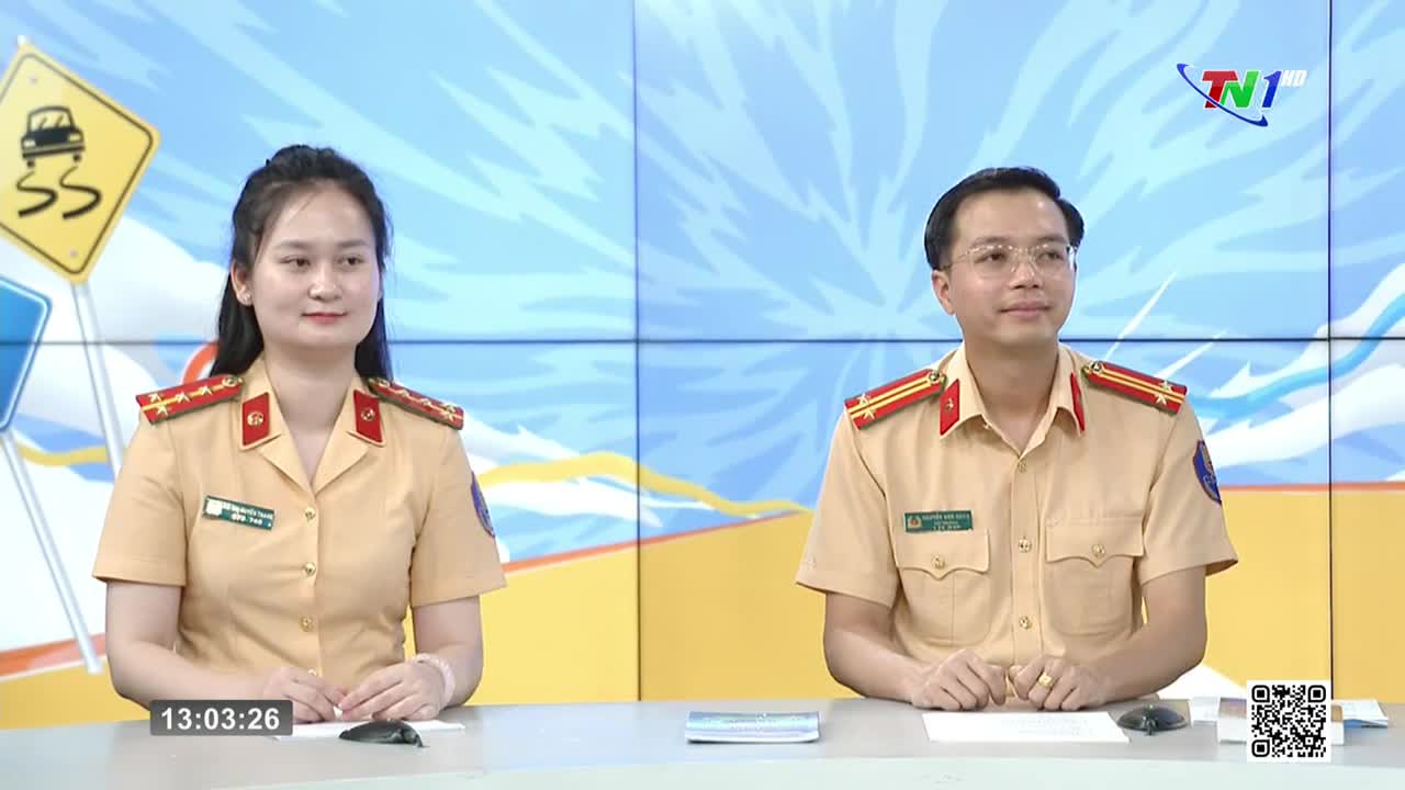 Two uniformed individuals sit at a desk, facing forward, with a Thai Nguyen TV logo visible in the background. The woman on the left has a small, pink bracelet, and the man on the right wears glasses.
