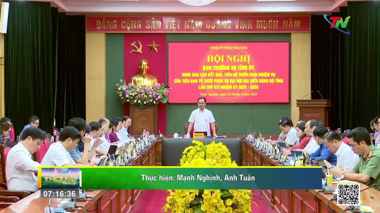 A man in a white shirt stands at a podium, addressing a room full of people seated at a long table. The scene is being broadcast on Thai Nguyen TV, the screen behind him displaying the meeting's agenda.
