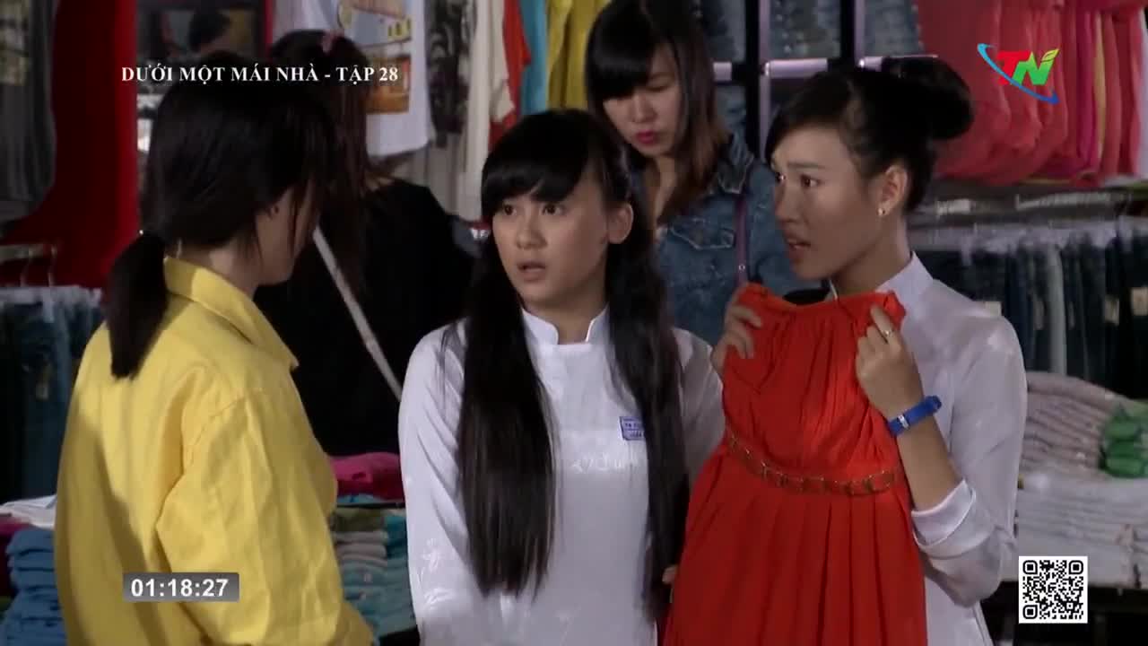 A woman in white holds up a bright red dress, gesturing animatedly to the others. The scene unfolds on Thai Nguyen TV, as the episode of "Duoi Mot Mai Nha" continues.
